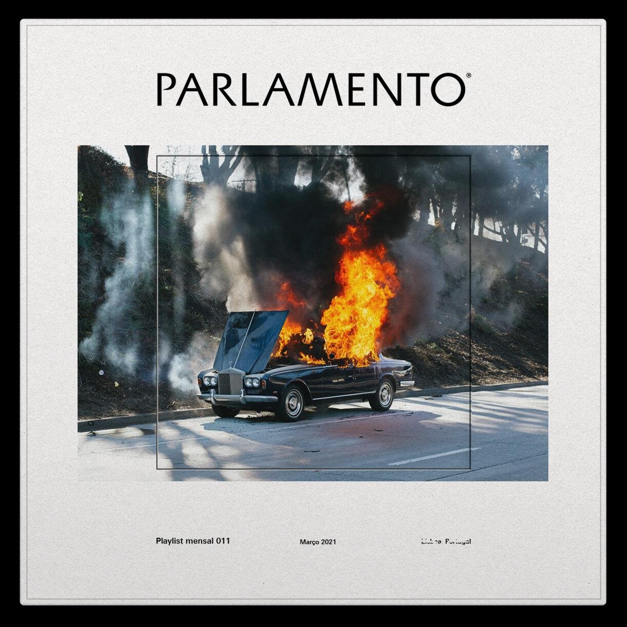 A vintage car is engulfed in flames on a road, with dark smoke rising above it.